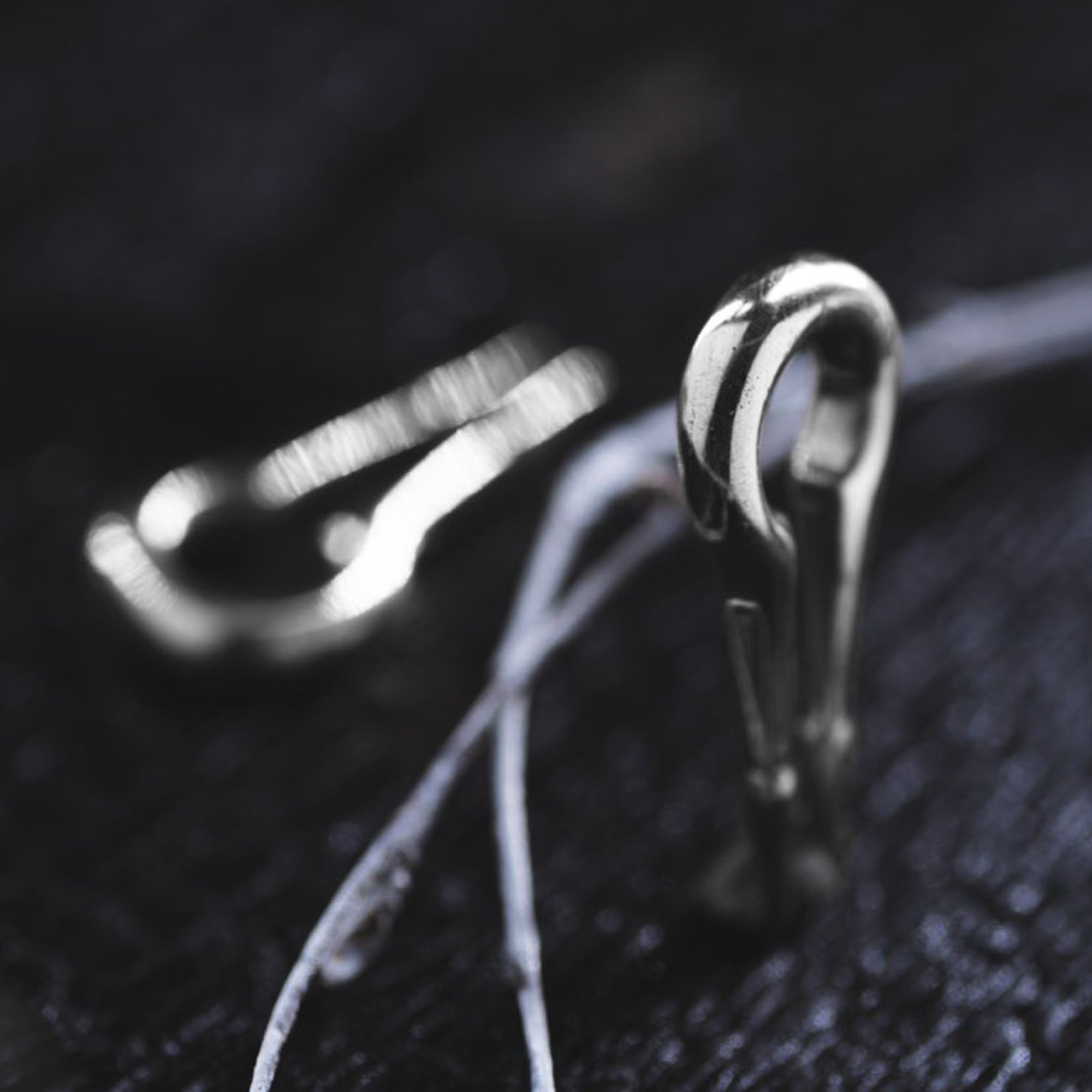 Close-up of a metallic carabiner clip lying on a dark surface. The clip is partially open, revealing its sturdy design. In the background, another blurry carabiner clip is visible, reminiscent of the CALYPSO Minimalist Keyhole Ear Weights in 925 Silver by Kaya Movement. Thin, light-colored wires or strings are scattered around them.